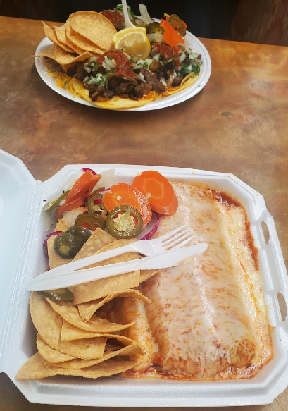 Authentic Mexican wet burrito with cheesy topping served with chips and pickled jalapeños, carrots, and onions from Taqueria Los Compadres, with a background plate of carne asada tacos.