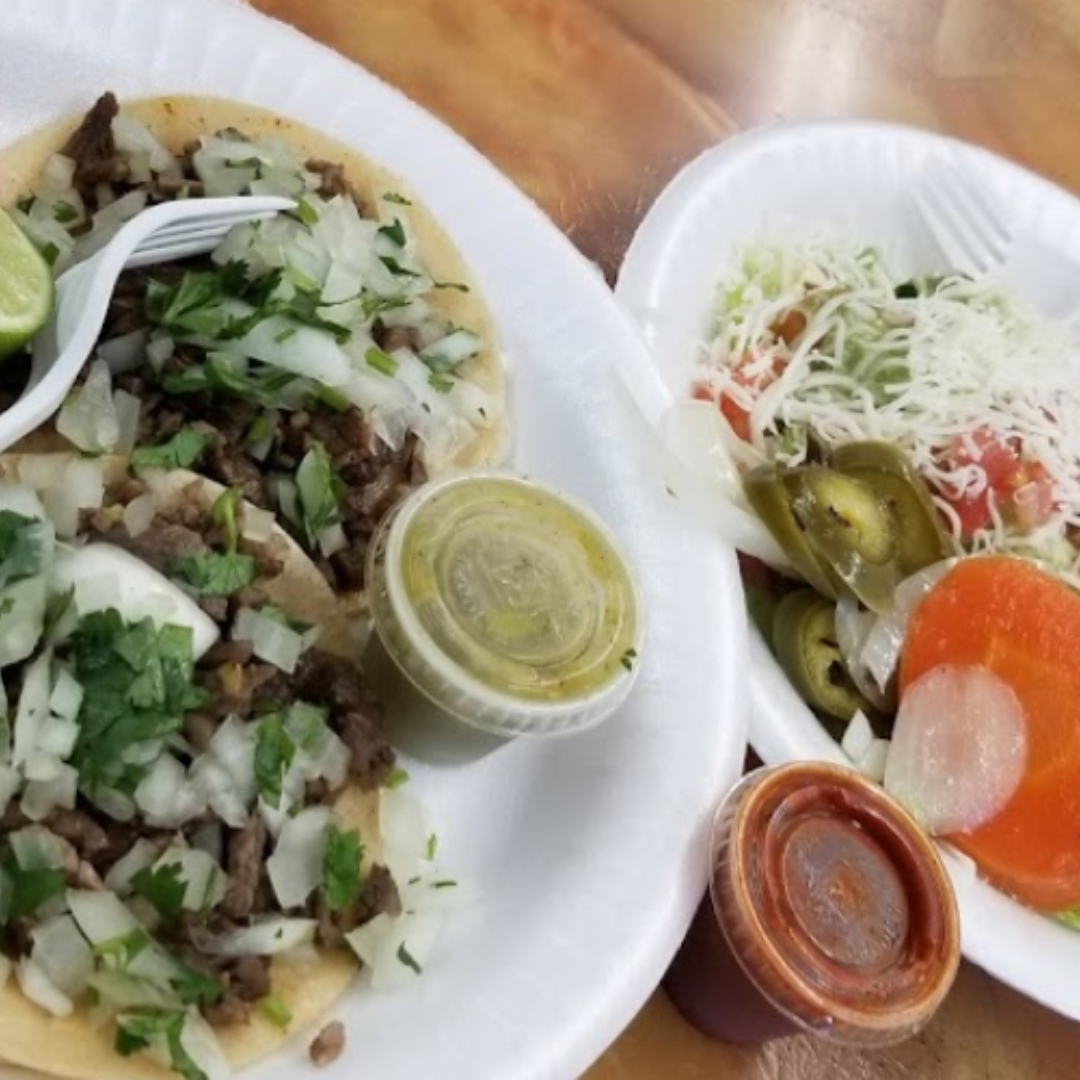 Savory carne asada taco with fresh cilantro and onions, accompanied by salsas and pickled jalapeños served on a foam plate at Taqueria Los Compadres.